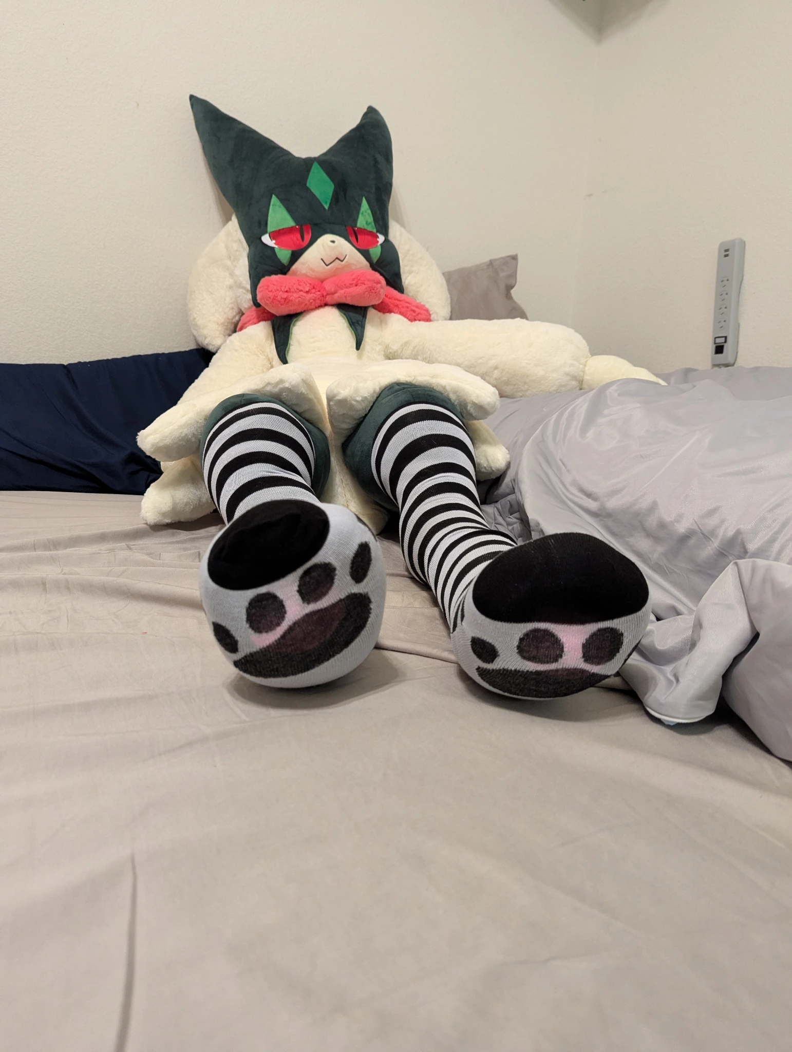 a photo of a meowscarada plushie laying on a bed, the meowscarada plushie has black and white striped thigh highs on its legs, there are cat paw prints printed on the bottom of the thigh highs.