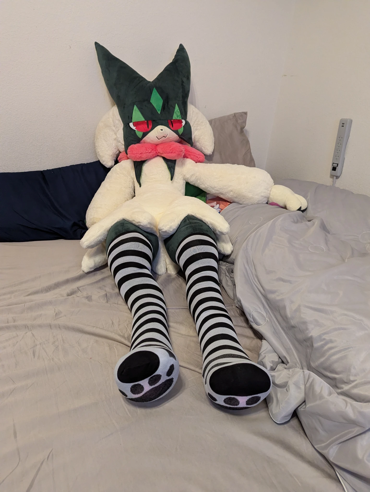a photo of a meowscarada plushie laying on a bed, the meowscarada plushie has black and white striped thigh highs on its legs, there are cat paw prints printed on the bottom of the thigh highs.